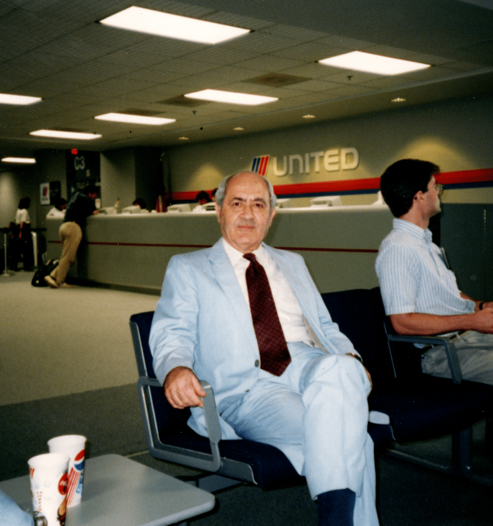 Dr. Naim Atiyeh in a powder blue suit with maroon tie relaxing in a chair with legs crossed in an airport lounge by the United Airlines gate.
