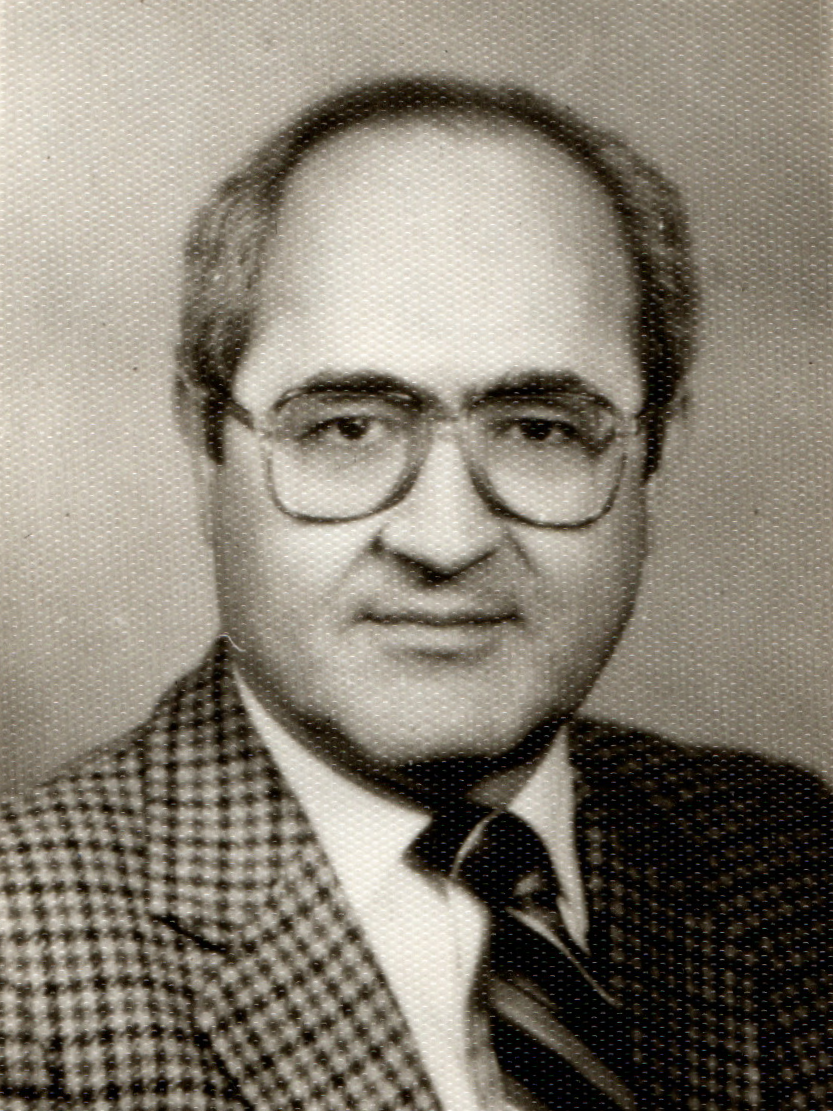 A black and white headshot of Dr. Naim Atiyeh wearing a square striped blazer, tie, and glasses.